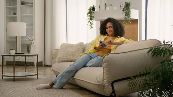 African American girl smiling young woman holding mobile phone using modern device on couch at home scrolling smartphone read social network laughing female lady blogger gadget addicted buying online