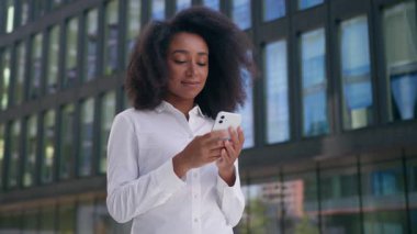 Afro-Amerikalı kadın internet üzerinden mesajlaşan dijital cihaz cep telefonu akıllı telefon sosyal medya kullanarak kameraya bakarak gülümse bayan iş kadını açık hava işvereni ile mesajlaşıyor.