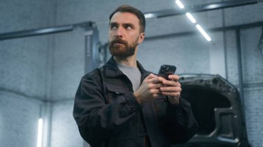 Tamirci tamirci. Otomobil tamirhanesinde cep telefonu aygıtı kullanıyor. Düşünceli erkek tamirci. Akıllı telefon ekranına bak.
