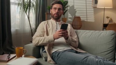 Düşünceli rüyalı beyaz erkek ev sahibi serbest kiracı evinde cep telefonu uygulaması kullanıyor. Apartman dairesinde internetten alışveriş yapıyor. Kanepede rahat ol.