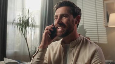 Kafkasyalı bir adam gülümsüyor mutlu, erkek, ev sahibi, kiracı, iş adamı, serbest çalışan, akıllı telefonu arıyor, cep telefonuyla konuşuyor, arkadaşça sohbet ediyor, kanepede rahat rahat sohbet ediyor.