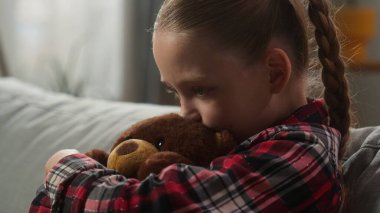 Stresli üzgün, hayal kırıklığına uğramış, yalnız, küçük, beyaz bir kız çocuğu evlat edinmiş, elinde oyuncak ayıcığıyla kanepeye oturmuş, aile içi şiddet, aile içi şiddet ve boşanma sorunları yaşıyor.