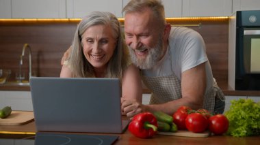 Olgun orta yaşlı, mutlu, beyaz aile çiftleri internetten yemek alışverişi yapıyor. Mutfakta dizüstü bilgisayarla yeni ürünler alıyorlar.