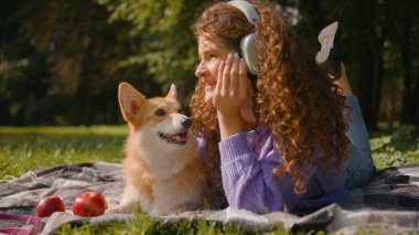 Mutlu Kafkasyalı kadın hafta sonu açık havada müzik kulaklığı dinliyor. Güzel bir köpek yavrusu yetiştiriyor. Çimenlerde tatlı köpek yavrusu yalıyor.