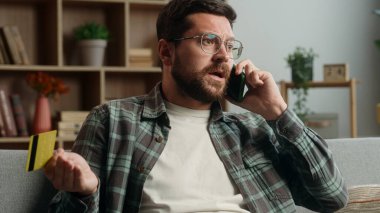 Stresli, sinirli, sinirli, Kafkasyalı erkek, online alışveriş yapıyor, çağrı desteği konuşuyor, banka konuşmalarına yardım ediyor, cep telefonu, akıllı telefon, evde kredi kartını bloke ediyor.