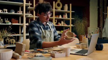 Erkek çömlekçi Kafkasyalı adam seramik stüdyosunda kil yontuyor dizüstü bilgisayarla el yapımı sanat eserleri modelliyor çevrimiçi video rehberliği yapıyor çömlekçilik atölyesindeki sanat atölyesinde öğretmenlik yapıyor.