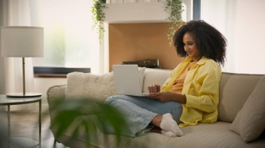 Afro-Amerikalı kadın ev sahibi kanepesinde dizüstü bilgisayarla gevşiyor internette sohbet ediyor bilgisayarla evde etnik kız iş kadını serbest meslek öğrenimi yapıyor.