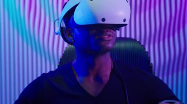 Sanal oyuncular, VR gözlüklü Afrikalı Amerikalı adam, kapalı mekanlarda, karanlık mavi odada sanal gerçeklik oyunu oynuyor. Erkek, siber-punk, moda oyunları yeniliği, 3 boyutlu simülasyon miğferi kullanıyor.