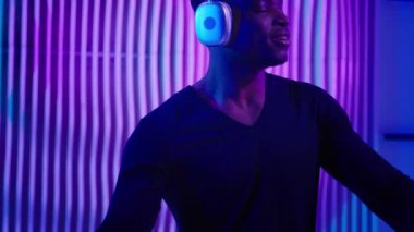 Afrika kökenli Amerikalı erkek dansçı erkek DJ modern kablosuz kulaklıklarda ses müziği dinliyor ev partisinde neon ışıklı modern hareketlerle eğleniyor özgürce hareket ediyor