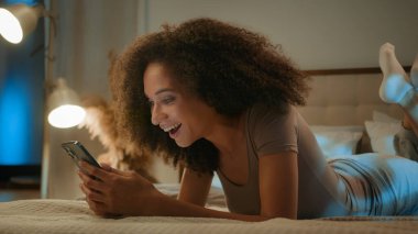 Afro-Amerikalı kadın mutlu bir şekilde genç kız cep telefonu kullanıcısı yatak odasında yatarken afallamış bir kadının akıllı telefon zaferini kazanmasına şaşırmış.