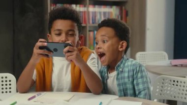 Okul eğitimi küçük çocuklar Afro-Amerikan kardeşler mutlu çocuklar çevrimiçi video oyunu oynuyorlar akıllı telefon bağımlısı iki öğrenci birlikte eğleniyorlar cep telefonu oyunu oynuyorlar.
