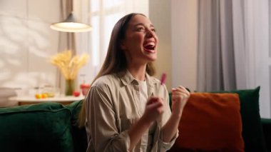 Hamilelik testi olan mutlu, beyaz bir kadın bebek sağlığı hekimliği için seviniyor. Başarılı bir kadın hamile. Kanepe üzerinde hamilelik testi yapıyor.