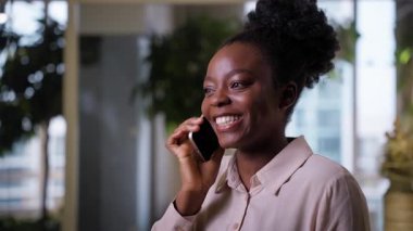 Ofiste cep telefonundan konuşan iş kadını mutlu, Afro-Amerikan, etnik, neşeli iş kadını, ofis içi konuşan, gülümseyen kız, akıllı telefon, kahkahalar, sohbet konuşmaları.