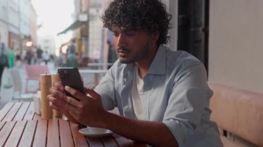 Kafe 'deki Hintli erkek Arap erkek öğrenci turist müşteri kahve içer düşünceli iş adamı seyahat bileti uygulamasını seçer internet siparişini internetten alışveriş cep telefonunu açık havada alır.