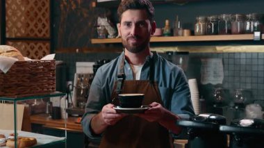 Gülümseyen beyaz erkek küçük işletme sahibi üniformalı kahve barmeni kapuçino barmeni işvereni kafeteryada çalışan müşterilere kahve dağıtıyor.