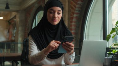 Müslüman kadın tesettüre girmiş Arap iş kadını. Masa başında çalışan gülümseyen iş kadını. Cep telefonu bağımlısı.
