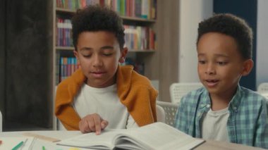 Afrikalı Amerikalı küçük çocuklar, kardeşler, öğrenciler, çocuklar, masa başında okuyan ilkokul öğrencileri, kütüphanede kitap okuyan öğrenciler, kameraya gülümseyen şirin yüzlere bakarak ders alın.
