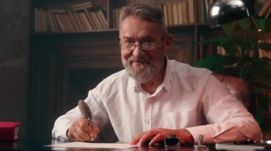 Başarılı beyaz adam elinde dolmakalem kâğıdı tutarken yazar, metin işadamı, kameraya bakıp gülümseyen emekli, ev içi ofis portresi çektiren olgun bir emekli.