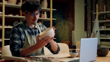 Erkek esnaf çömlekçi, sanat stüdyosunda kil yontuyor. Kafkasyalı adam odaklı erkek tasarımcı, tasarım tasarımcısı. Elinde çanak gibi el işi el işi sanat eseri, çömlekçilik atölyesinde seramik boyuyor.