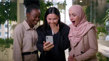 Çok ırklı kızlar, Afrikalı Müslüman Asyalı iş arkadaşları iş arkadaşları mutlu neşeli kadınlar cep telefonu uygulaması kullanarak zaferi kutluyorlar.