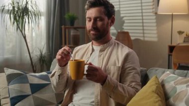 Beyaz erkek, ev sahibi, erkek, kanepede oturuyor, kahve içiyor, dinleniyor, elinde bardakta sağlıklı içecek, ilaç alıyor, sağlık hizmetleri, ilaç tedavisi, içecek, rahatlama, gülümseme, evde rahatlama.