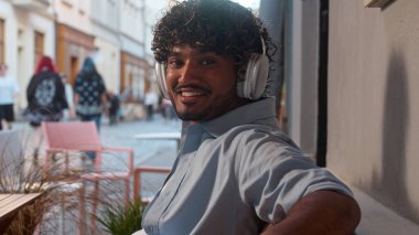 Arap Hindistanlı mutlu adam erkek turist Kafe restoranında ziyaretçi açık havada kahve iç rahatla kulaklıklarda müzik dinle neşeli gülümseme kameranın ritmine bak