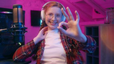Kadın blogger vlogger etkileyicisi radyo podcast mikrofon akışı kulaklıklarla Kafkasyalı genç kız bilgisayar oyunu siber oyun bağımlısı, kamera jestine bakıyor.