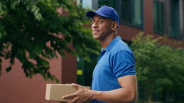 Postacının kaygısız erkek çalışanı postacı kurye adam gülümsüyor karton kutu standında çalışıyor şehir e-ticaret alışveriş postacılığı sokak kameralarına bakıyor.