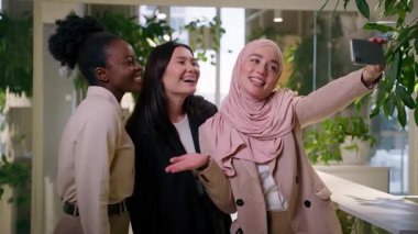 Ofisteki üç farklı çok ırklı iş kadını gülümseyen mutlu kızlar cep telefonu kamerası öğrencilerine poz veren Asyalı Müslüman Afrikalı iş kadınları iş arkadaşları neşeli kadın selfie akıllı telefon sabıkası medyası