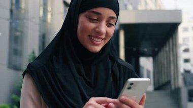 Mutlu gülümseyen Müslüman kadın tesettür giyiyor iş kadını şehir merkezindeki iş kadını cep telefonu gülümsemesi kullanıyor kaygısız sosyal medya akıllı telefon uygulamaları kullanıyor