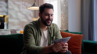 Gülümseyen mutlu beyaz sakallı erkek ev sahibi gülen model iş adamı kameraya bakıyor yığınla para kazanıyor kumar oynuyor zafer ödülü kazanan ev sahibi oturma odasında piyango kazanıyor