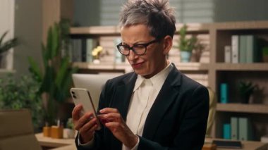 Kafkasyalı iş kadını yaşlı Avrupalı iş kadını olgun, olgun, kadın, emekli, iş adamı. Ofisteki işveren, cep telefonlu, hoşnutsuz, akıllı telefon kullanan, kötü haber okuyan, somurtkan, duygulu.