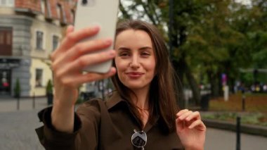 Gülümseyen Kafkasyalı kadın iş kadını şehir caddesinde elinde cep telefonu, telefon konferansı, video kaydı, fotoğraf çekimi, el sallama.