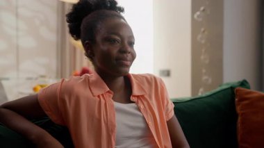 Rahatlamış, gülümseyen Afro-Amerikan etnik kadın mutlu bayan model ev sahibi gülen ev sahibi müşterimiz TV film komedisi seyrediyor kanepede kanepede oturuyor hafta sonu boş vakitlerinde evde oturma odasında