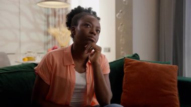Düşünceli, düşünceli, Afro-Amerikan etnik kadın şaşkın kadın model ev sahibi müşteri çeneye dokunmayı düşünüyor derin düşüncelere dalmak problem çözümünü evde oturma odasında kanepede oturarak çözmek