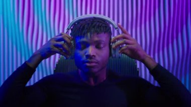 Afro-Amerikalı etnik bir adam kulaklık takmış kulaklık takıyor kulaklık takmış müzik dinliyor müzik dinliyor internet müzik dinliyor modern kulaklıkla neon oda ortamında fütüristik titreşimler yayıyor