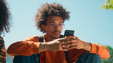 İki Afrikalı Amerikalı öğrenci, kız arkadaş erkek erkek erkek erkek, alet bağımlısı cep telefonları, akıllı telefonlar sosyal medya, üniversite üniversitesinin açık hava yerinde oturuyorlar.