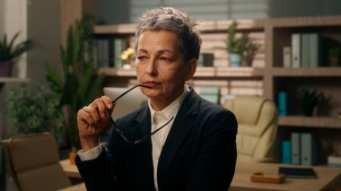 Ofiste düşünceli, yaşlı, düşünceli, beyaz kadın, iş kadını, olgun, orta yaşlı, elinde gözlükleriyle gülümseyen bir işveren, kameraya bakarak bir fikir üretiyor.