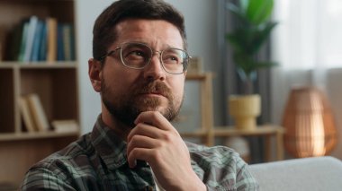 Düşünceli, düşünceli, tereddütlü, kararsız, düşünceli, kafası karışmış, kafası karışmış, kafatası karışık bir erkek evin içinde kameraya gülümseyen yaratıcı çözümler bulmaya çalışıyor.