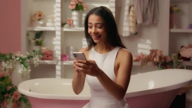 Şeker banyosundaki mutlu Latin kadın neşeli kız çevrimiçi bahsi kazanmayı kutluyor cep telefonu akıllı telefon başarısını kullanarak zafere ulaşıyor.