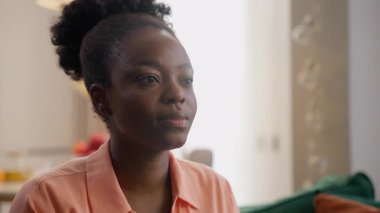 Sakin, ciddi, düşünceli, düşünceli, Afro-Amerikan iş kadını, etnik bayan, model, müşteri, ev sahibi, ev sahibi, ev sahibi, kameraya bakıyor, kanepede, evde oturma odasında oturuyor.