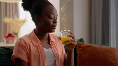Rahatlamış, mutlu, sağlıklı, gülen, Afro-Amerikan, iş kadını, bayan, model, müşteri, ev sahibi, TV seyreden ev sahibi, taze portakal suyu içen, içecek, oturma odası içeceği içen kadın.
