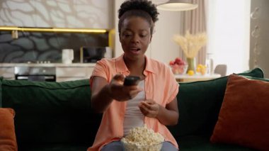 Mutlu kahkahalar atan Afro-Amerikan iş kadını ev sahibi bayan bayan iş kadını rahatla kanepe seç TV kumandanı tut patlamış mısır ye evde film izle televizyon programı izle