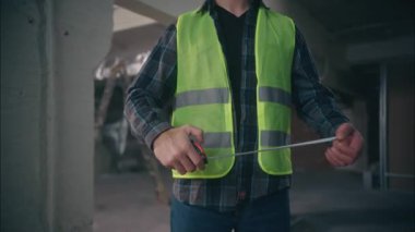 Kasklı, kasklı beyaz bir adam. Rulet aletlerini ölçmeye bakıyor. Müteahhit mühendis.