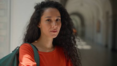 Kafkasyalı, ciddi, barışçıl, Latin kökenli, Latin kökenli, bayan öğrenci üniversite akademisi koridorunda eğitim öğrenim görüyor ve kamera fotoğraflarına bakıyor.