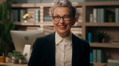 Ofisteki mutlu, gülümseyen dişlek, sağlıklı gülümseyen yaşlı, gri saçlı, olgun, yaşlı iş kadını, kadın, işveren, menopoz tıbbı kameraya bakıyor.
