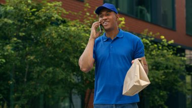 Afro-Amerikalı kurye konuşuyor. Müşteri cep telefonunu arıyor. Elinde kağıt paketler, yiyecek teslimatı postacı, erkek karton teslimat.
