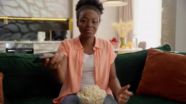 Kafası karışık, mutsuz, Afro-Amerikan kadın, etnik kız, kullanıcı kontrolörü, kase patlamış mısır yiyor, kanal arızasını değiştirmeye çalışıyor.