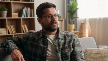 Evde mutlu, sakallı, mutlu bir erkek portresi. Ev kredisi sigortası. Kanepede rahatlayıp kamera karşısında gülümseyen, dişlek, diş hekimi gözlüğü takan adam.