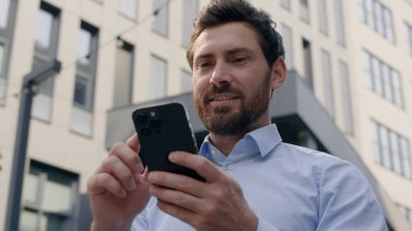 gülümseyen Kafkasyalı orta yaşlı iş adamı erkek işadamı işveren iş adamı şehirdeki ofis binası yakınında cep telefonu uygulaması kullanarak akıllı telefon sohbeti yapan akıllı telefon işi internet kullanan girişimci